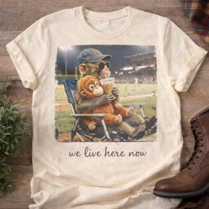 May include: A cream-coloured t-shirt with a graphic print of two monkeys at a baseball game. One monkey wears sunglasses and a cap, holding a drink, while the other is an orange plush toy. The text "we live here now" is printed below the image.