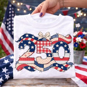 May include: White t-shirt with a patchwork design of the number "250", incorporating red, white, and blue patterns. The design includes stars, stripes, and a bow, with a patriotic theme. The shirt is held up in front of an American flag.