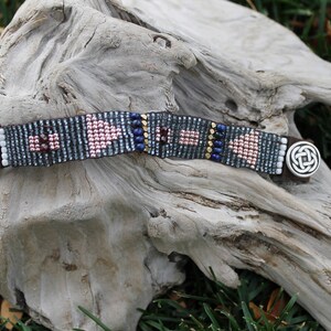 May include: A beaded bracelet with a brown leather strap and a silver clasp. The beads are in shades of blue, pink, and gold, and are arranged in a geometric pattern.