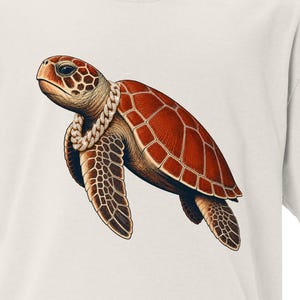 May include: A cream-colored short-sleeve t-shirt featuring a graphic illustration of a sea turtle wearing a thick silver-toned chain necklace. The turtle has a detailed reddish-brown shell with distinct scutes and patterned flippers.
