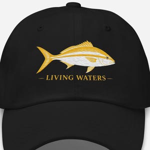 May include: A black baseball cap featuring a graphic of a yellow and white fish centered on the front panel. Below the fish, the text "LIVING WATERS" is printed in a yellow serif font, flanked by two horizontal lines on either side of the text.
