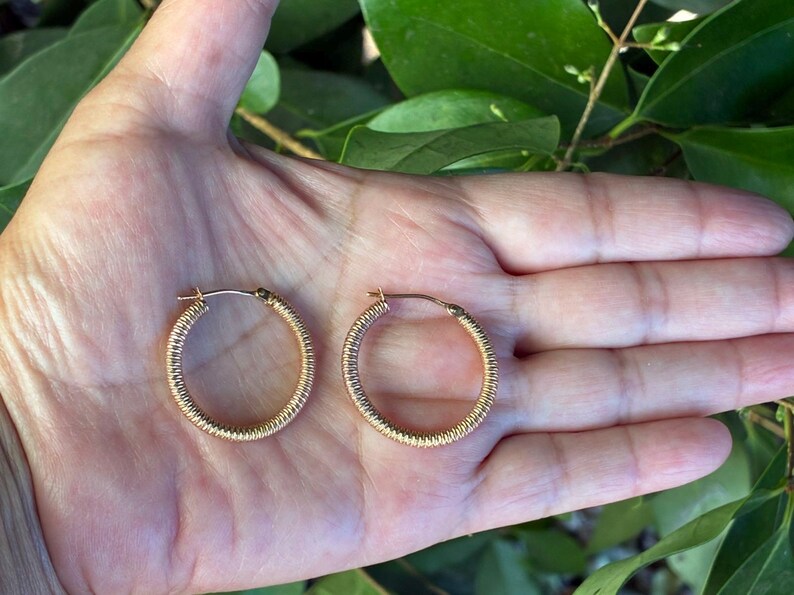 May include: A pair of gold-colored hoop earrings with a textured, ridged design. The earrings are circular and appear to be made of metal. The earrings are displayed on an open palm against a background of green leaves.