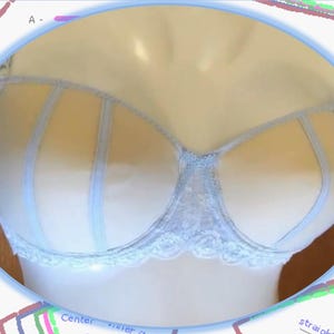 Puede incluir: Un sujetador de encaje azul claro con un diseño delicado. El sujetador tiene una copa transparente con líneas verticales y un borde de encaje.