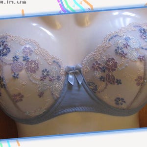 以下が含まれることがあります： 紫と青の花柄刺繍が施された、淡いブルーのブラジャー。ブラジャーは、上端に繊細なレースのトリムと中央に小さなリボンが付いています。マネキンに展示されています。