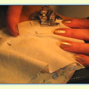 May include: Close-up of a sewing machine needle going through a piece of white fabric. The fabric has markings on it, and the person's fingers are visible on the fabric.