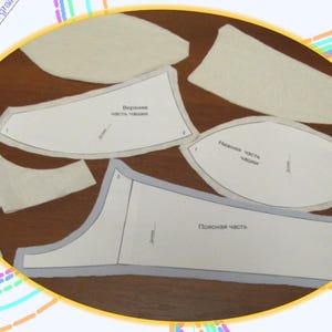 May include: A set of white fabric patterns for sewing a bra. The patterns are labeled in Russian. The patterns include pieces for the upper cup, lower cup, and side panels.