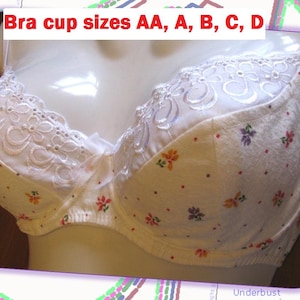Puede incluir: Un sujetador blanco con un delicado estampado floral de pequeñas flores rojas, naranjas y moradas, con un borde de encaje blanco bordado en la parte superior. El texto "Bra cup sizes AA, A, B, C, D" aparece impreso en rojo en la parte superior.