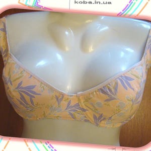 May include: A peach-colored bra with a floral pattern of lavender and white leaves. The bra is displayed on a mannequin. The bra has adjustable straps and a white trim along the neckline.