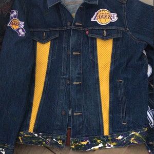 May include: A blue denim jacket with yellow and white accents. The jacket has two patches on the front, one with the Los Angeles Lakers logo and the other with the text "Los Angeles Lakers".
