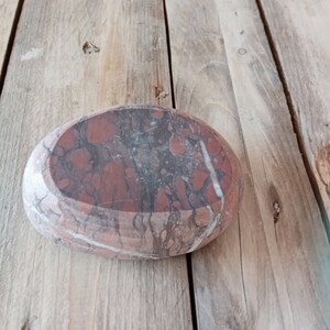 Può includere: Primo piano di una pietra ovale rossa e bianca con un motivo marmoreo. La pietra è appoggiata su una superficie di legno.
