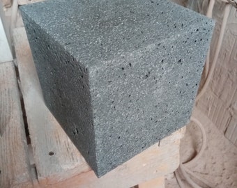 May include: A dark gray, rough-textured, square-shaped block of volcanic rock. The block is sitting on a wooden surface.