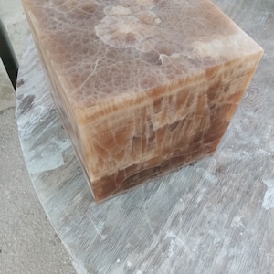 Pode incluir: Um pedestal quadrado de ônix de cor marrom claro com um padrão veteado único. O pedestal é feito de pedra natural e tem um acabamento liso e polido.