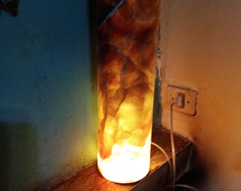 May include: A cylindrical lamp made of translucent, brown and yellow onyx with a warm, glowing light emanating from inside. The lamp is sitting on a wooden shelf.