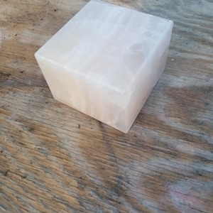 Pode incluir: Uma caixa decorativa quadrada branca feita de cristal de selenite. A caixa tem uma superfície lisa e polida e está sentada em uma superfície de madeira.