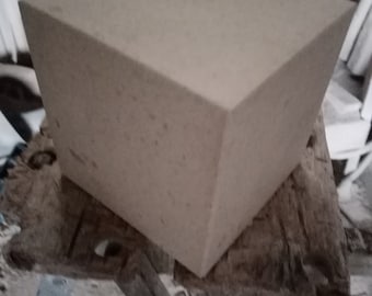 May include: A large, square, light beige block of stone. The block is sitting on a rough, wooden surface.