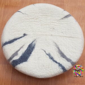 Op de afbeelding: Een rond, wit, gevoeld kussen met een zwart en grijs marmerpatroon. Het kussen is gemaakt van wol en is perfect om elke kamer een vleugje textuur en kleur te geven.