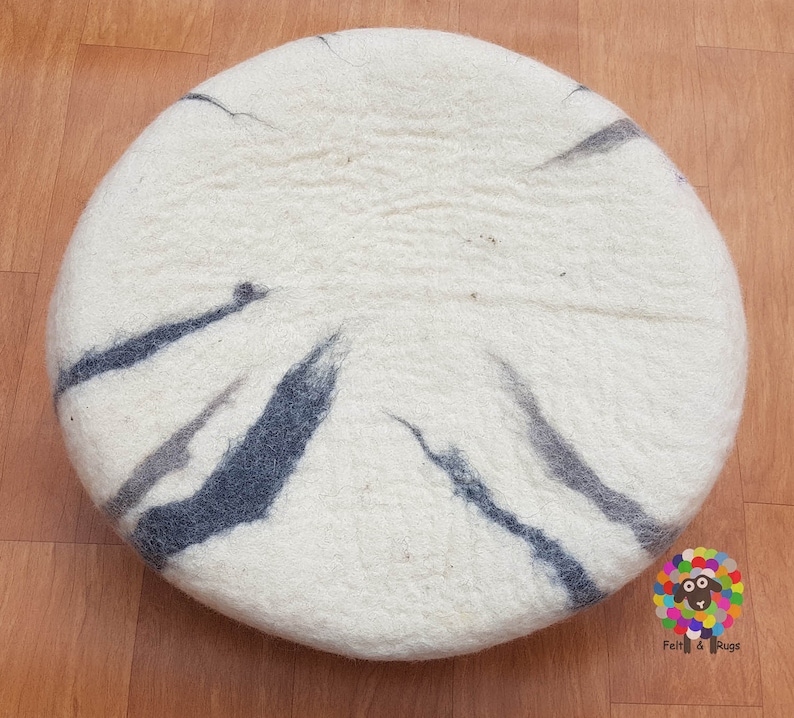 Op de afbeelding: Een rond, wit, gevoeld kussen met een zwart en grijs marmerpatroon. Het kussen is gemaakt van wol en is perfect om elke kamer een vleugje textuur en kleur te geven.