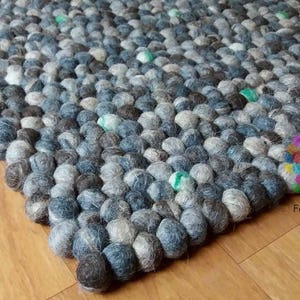 May include: A close-up of a grey and white wool rug made of small, round balls. The rug is made by Felt & Rugs.