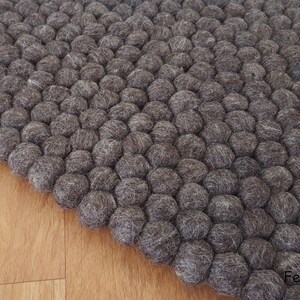 May include: A close-up of a gray felt ball rug. The rug is made of many small, round felt balls, creating a textured surface. The rug is on a light brown wooden floor. The logo "FeltAndRugs" is in the bottom right corner.