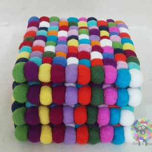Puede incluir: Un posavasos de fieltro colorido hecho de pequeñas bolas de fieltro en varios colores, incluyendo rojo, verde, azul, amarillo, morado y blanco. El posavasos está hecho de múltiples capas de bolas de fieltro, creando un diseño grueso y resistente.