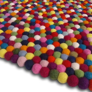 Puede incluir: Una alfombra colorida hecha de bolas de fieltro en varios tonos de rojo, naranja, amarillo, verde, azul, morado, rosa y blanco. Las bolas están dispuestas en un patrón escalonado, creando una superficie texturizada y visualmente atractiva.