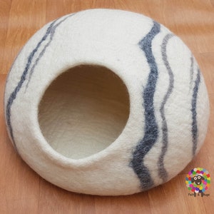 Pode incluir: Cama redonda para gatos de feltro branco com uma grande abertura. A cama tem um padrão ondulado de feltro cinza escuro e cinza claro.