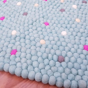 Könnte beinhalten: Ein runder Filzteppich in hellblau mit weißen, rosa und grauen Filzkugeln. Der Teppich ist aus Wolle und perfekt für ein Kinderzimmer oder einen Spielbereich.