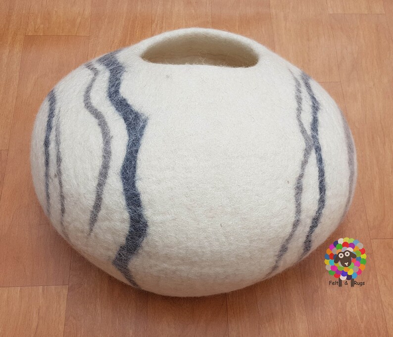 Op de afbeelding: Rond kattenbed van wit vilt met een gat om in te gaan. Het bed heeft een grijs en blauw gestreept patroon.