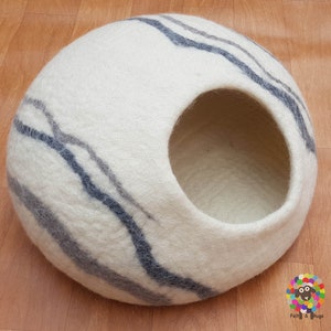 Op de afbeelding: Een wit wollen kattenbed met een grijs en wit gestreept patroon. Het bed heeft een ronde opening waar de kat in kan.