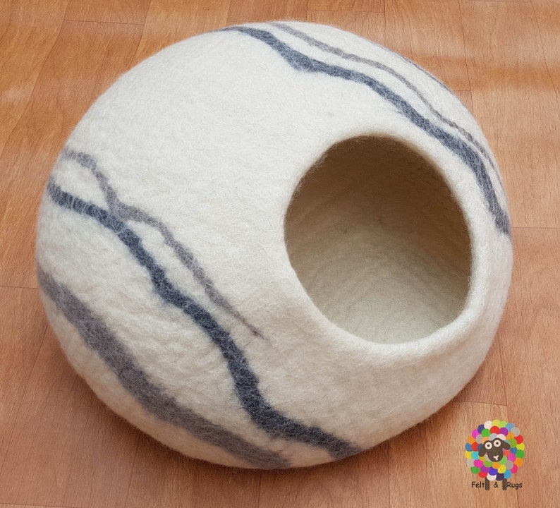 Op de afbeelding: Een wit wollen kattenbed met een grijs en wit gestreept patroon. Het bed heeft een ronde opening waar de kat in kan.