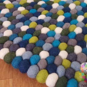 Pode incluir: Um tapete redondo de feltro colorido feito de pequenas bolas densamente embaladas. O tapete é feito em uma variedade de tons de azul, verde e branco.
