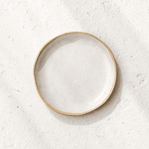 Puede incluir: Un plato de cerámica blanco con un esmalte moteado y un borde beige. El plato está sobre una superficie blanca texturizada.