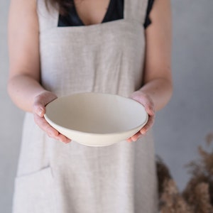 Può includere: Una persona che indossa un grembiule beige chiaro tiene una ciotola di ceramica bianca nelle sue mani.