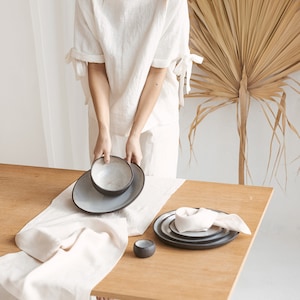 May include: A white linen table runner with a white linen napkin and a set of black and white ceramic bowls and plates on a wooden table.