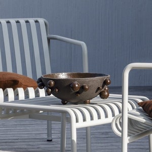 Puede incluir: Dos sillas de metal blanco con cojines marrones y una mesa de metal blanco con un cuenco marrón en la parte superior. Las sillas y la mesa están en una terraza de madera frente a una pared azul.
