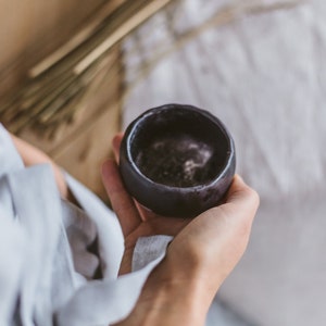 Op de afbeelding: Een kleine, donkergrijze keramische kom met een licht getextureerd oppervlak. De kom is leeg en lijkt handgemaakt te zijn.