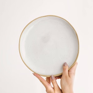 Può includere: Un piatto in ceramica bianca con una glassa macchiettata e un bordo beige. Il piatto è tenuto da due mani.