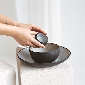 Puede incluir: Un juego de tres cuencos de cerámica negra, uno pequeño, uno mediano y uno grande. Los cuencos están anidados uno dentro del otro y están sobre una superficie blanca texturizada.