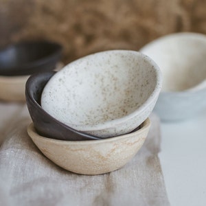 Può includere: Un set di quattro piccole ciotole in ceramica in varie tonalità di beige, marrone e bianco. Le ciotole sono punteggiate da piccole macchie marroni e hanno una superficie leggermente ruvida.