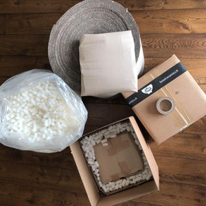 May include: Overhead shot of packaging materials on a wooden floor. Includes a roll of woven fabric, a wrapped package, a box with tape and a logo, and a clear bag of packing peanuts. A second box is open, filled with packing peanuts.