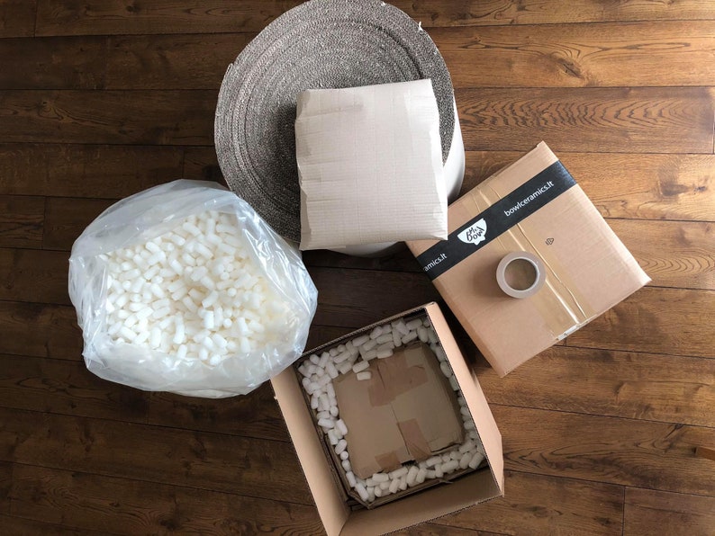 May include: Overhead shot of packaging materials on a wooden floor. Includes a roll of woven fabric, a wrapped package, a box with tape and a logo, and a clear bag of packing peanuts. A second box is open, filled with packing peanuts.