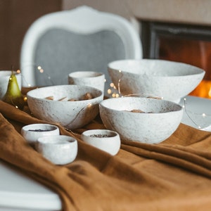 Può includere: Un set di cinque ciotole in ceramica bianca con smalto grigio macchiato. Le ciotole sono disposte su una tovaglia marrone con un tavolo bianco sullo sfondo. Le ciotole sono di diverse dimensioni e sono perfette per servire spuntini o dessert.
