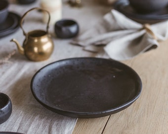 May include: A black ceramic plate with a slightly textured surface sits on a wooden table. The plate is empty and ready for a meal.