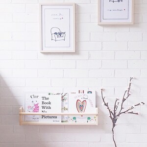 May include: A wooden bookshelf, mounted on a white brick wall, displays children's books. Above, two framed prints show a person in a chair with the text "ahh, I'm reading" and "my sundays". A decorative branch is in a pink basket.