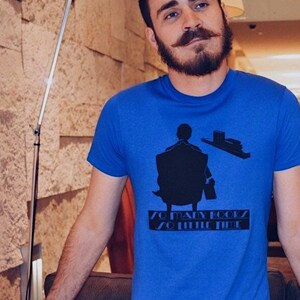 K&ouml;nnte beinhalten: Ein blaues T-Shirt mit einer schwarzen Grafik einer Person, die in einem Stuhl sitzt, einem Stapel B&uuml;cher und dem Text "SO MANY BOOKS SO LITTLE TIME". Das Shirt ist aus einem weichen Material.