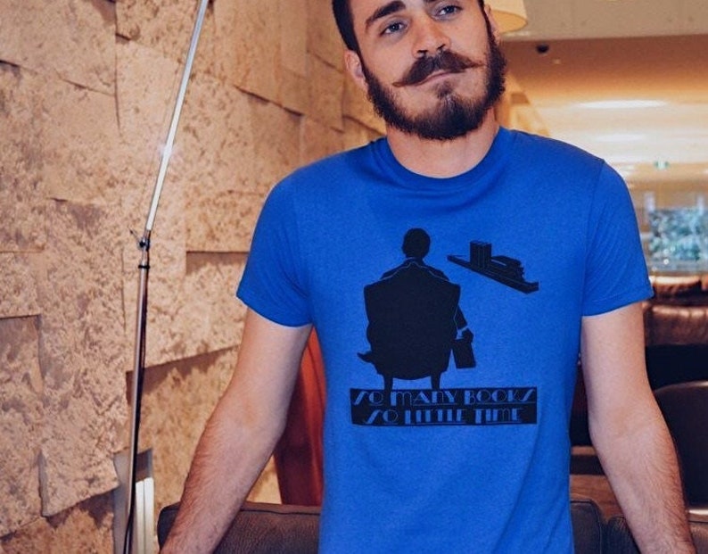 K&ouml;nnte beinhalten: Ein blaues T-Shirt mit einer schwarzen Grafik einer Person, die in einem Stuhl sitzt, einem Stapel B&uuml;cher und dem Text "SO MANY BOOKS SO LITTLE TIME". Das Shirt ist aus einem weichen Material.