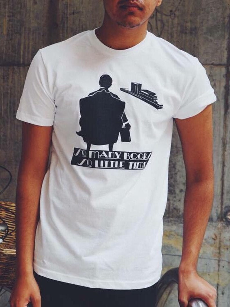 K&ouml;nnte beinhalten: Wei&szlig;es T-Shirt mit schwarzer Grafik einer Person, die in einem Stuhl sitzt, mit B&uuml;chern und dem Text "So many books, so little time". Das Shirt besteht aus weichem Material und hat kurze &Auml;rmel.