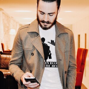 K&ouml;nnte beinhalten: Ein grauer, zweireihiger Mantel &uuml;ber einem wei&szlig;en T-Shirt mit schwarzer Grafik. Das T-Shirt zeigt eine Silhouette einer Person und den Text "MY BOOK LITTLE TIME". Die Person h&auml;lt ein schwarzes Telefon.