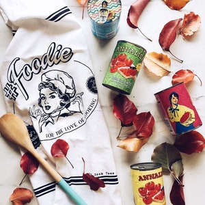 May include: A white kitchen towel with the word "Foodie" and a vintage illustration of a chef. Several cans of tomatoes and other food items are scattered around the towel, along with a wooden spoon and autumn leaves. The towel reads "For the love of cooking."