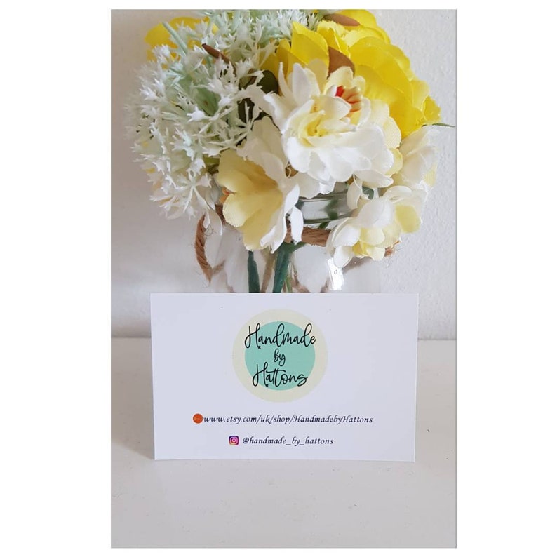 May include: A white card with the text "Handmade by Hattons" in a circle with a teal background. The text "www.etsy.com/uk/shop/HandmadebyHattons" and "@handmade_by_hattons" are below the circle. The card is in front of a bouquet of white and yellow flowers in a glass jar.